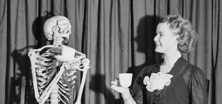 Black and white photo from the 1930's featuring a smiling young lady having tea with a skeleton.